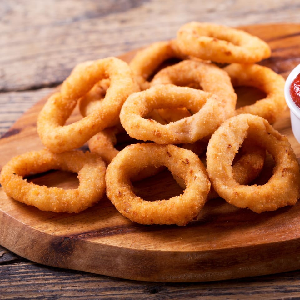 Golden-brown onion rings piled on a wooden board, with a side of ketchup, at JD's Grill House.