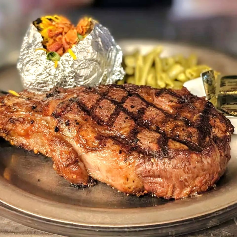 Grilled steak with a foil-wrapped baked potato and green beans on a metal plate at JD's Grill House.