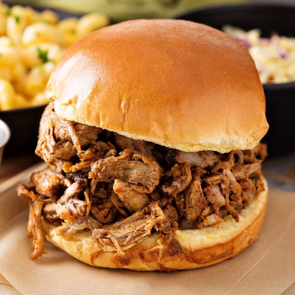 Close-up of a JD's Grill House pulled pork sandwich on a bun with sides in the background.